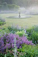 Dawn light on the fountain in the lawn with blue borders around. Narborough Hall Gardens, Norfolk