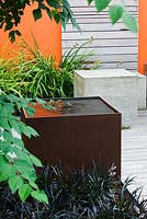 Modern contemporary garden in Brighton with square metal water feature, concrete seats on decking with wooden panel walls and orange panel 
