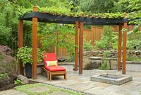 Summer garden with black steel and cedar arbor and red lounge chair. Hardscaping includes stone pavers, fire pit, waterfall, stacked stone wall, cedar fence. 
