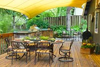 Summer outdoor kitchen under canopy with grill and sink on wooden deck. Wrought-iron chairs and table with succulent garland centerpiece.