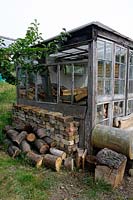 Allotment shed, hut or glasshouse made of stripped wood with wooden chaise lounge in front and with bricks stacked alongside and logs inside and outside. Golf Course Allotments, London Borough of Haringey.