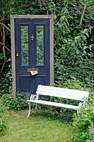 Victorian front door used as Trompe l'oeil in a garden in Hackney, with junk mail in letterbox and white painted bench to fore.