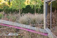 Decorative pink steel and wooden bridge over pond to summerhouse through Molinia 'Transparent' - Farrs
