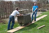 Men transporting granite trough over lawn using planks of wood