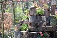 Wooden birdhouses, tulips in pots, planted fritellarias, and small beehotels on stacked tree trunks.