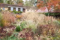 Gravel path leading into the central circular bed framed by miscanthus, rosemary, Stipa gigantea, salvias, Sedum 'Purple Emperor' and Calamagrostis brachytricha, with prunus turning orange against red oxide barn roof behind. Broughton Buildings, Broughton, nr Stockbridge, Hants, UK