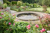 Circular pond on the terrace is surrounded by box edged beds containing roses including stripey Rosa 'Ferdinand Pichard' underplanted with hardy geraniums, poppies and alliums. Forest Lodge, Pen Selwood, Somerset, UK