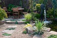 Pink gravelled area edged by boulders and stepping stones leading to circular patio with table and chairs and pond with water pump