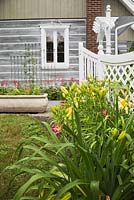 Partial view of a stacked log home with pink and yellow daylily (Hemerocallis) flowers and a white wooden fence with trellis frames - Il Etait Une Fois garden, Monteregie, Quebec, Canada. 
