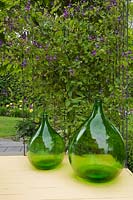 Two large green glass jugs next to a metal trellis with mauve Clematis flowers on a yellow painted wooden plank back porch - Il Etait Une Fois, Quebec, Canada.