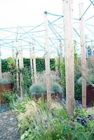 Several wooden piles connected with a blue rope standing in a border with Verbena Bonariensis. Rusted metal round containers planted with Pennisetum alopecuroides Hameln.