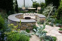 Hambrooks Halo garden  Mediterranean Greek island style. Rough-hewn stone paving with circular conversation seating with fire pit and table and chairs. Coastal seaside planting with verbasum. Sails in background. Designer: Stuart Charles Towner Silver-Gilt award.  