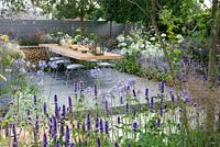 Vestra Wealth's Vista Garden. View through border with Agastache towards laid dining table for outdoor entertaining set on a grey-painted decking surrounded by blue palette planting with Agapanthus and hydrangea Annabelle and pleached hedging. Designer: Paul Martin Sponsor: Vestra Wealth Gold award  