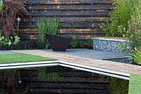 Elemental - reflective dark pool pond with charred burned redwood board wall basalt cobbles in cage with timber seating and path firepit - planting of grasses including Miscanthus sinensis 'Zebrius' zebra grass and bamboo Phyllostachys aurea 