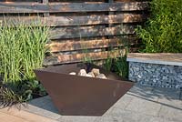 Firepit with charred burned redwood timber board wall - basalt patio stone and basalt cobbles in cage with timber seating, planting of grasses including Miscanthus sinensis 'Zebrius' zebra grass with  bamboo Phyllostachys aurea