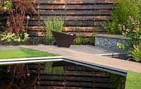 Elemental - reflective dark pool with charred burned redwood board wall basalt cobbles in cage with timber seating and path firepit - planting of grasses including Miscanthus sinensis 'Zebrius' zebra grass and bamboo Phyllostachys aurea