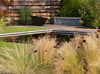 Elemental - reflective dark pool with charred burned redwood board wall, basalt cobbles in cage with timber seating and path firepit - planting of grasses including Miscanthus sinensis 'Zebrius' zebra grass and bamboo Phyllostachys aurea 