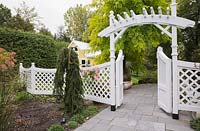 Grey flagstone path leading to a arbour through a white trellis fence in front garden in autumn. Plantings include Black locust, Robinia pseudoacacia 'Frisia' tree in the background. Il Etait Une Fois garden, Monteregie, Quebec, Canada. 
