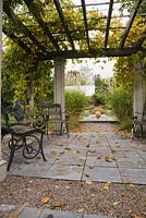 Wooden garden chairs and fallen leaves on flagstones underneath a wood and concrete pergola covered with Actinidia kolomikta 'Arctic Beauty' in backyard garden in autumn. Il Etait Une Fois garden, Monteregie, Quebec, Canada. 
