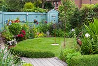 Cottage garden with circular lawn and planting which includes the marginal plant Calthra palustris by the small wildlife pond. Blue fence and garden shed.