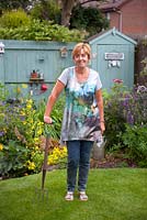 Garden designer, Angie Barker rests on garden fork in front of cottage garden border with Calthra palustris, Lupinus 'Gallery Blue' and small wild life pond