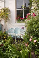 Decorative blue metal seat in colourful town garden with Geraniums in windowbox, Rosa 'Guinee', Papaver, Peonies Alliums and Centaurea
