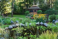 Pond with white Nymphaea alba - water lilies, Rudbeckia fulgida 'Goldsturm' - yellow coneflowers and a gazebo in the background in a backyard garden in summer, Laurentians, Quebec, Canada