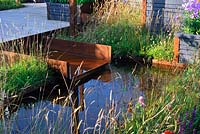Morning low light on grasses around shallow pool with rusty metal channel Waterfall. Description: Maggie's Forest Garden. Designer: Amanda Waring and Laura Arison. 