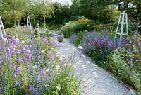 Gravel pathway thorugh flower garden planted with a mix of flowering herbaceous perennials with white wooden obelisks to give year round structure.