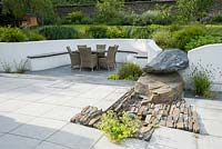 Contemporary terrace with rendered white retaining wall, naturalistic rock outcrop, dome fountain water feature and rattan garden furniture.