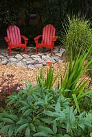 Two red wooden Adirondack chairs on  flagstone patio next to  Miscanthus sinensis 'Strictus', Paeonia lactiflora in the foreground and Berberis thunbergii in backyard garden in summer, Under the Apple Trees garden, Canada