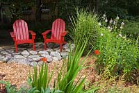 Two red wooden Adirondack chairs on flagstone patio next to Miscanthus sinensis 'Strictus' - Zebra Grass, pink flowering Physotegia virginiana - Obedient Plant, red Crocosmia montbretia 'Lucifer' 