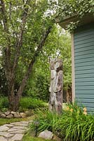 Jack-rabbit Johanssen wooden totem pole next to a garden border with orange Hemerocallis - Daylilies and a flagstone path in backyard garden in summer, Malus sylvestris - Wild Apple tree in border edged with stones