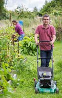 Man mowing the lawn in an allotment, woman working in the background