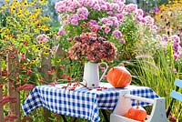 Table with harvested squashes 'Hokkaido' and jug of Hydrangea seedheads, rosehip and Verbena bonariensis. Border - Pennisetum 'Hammeln', Sedum, Rudbeckia triloba, Aster, Panicum 'Heavy Metal'.