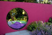 View through pink moon wall with bean bags in relaxing area. Planting includes Nepeta 'Six Hills Giant', Alchemilla mollis