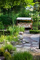 Swing seat in summer garden, Rosa 'Alchymist', Rosa 'Golden Showers', path, stone slabs used as stepping stones in gravel, pond
