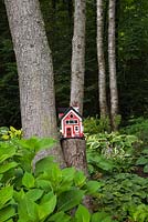 Red, white and black wooden birdhouse on a tree stump surrounded by hostas and pteridophyta - ferns woodland garden in summer