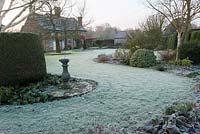 Frosty garden with mixed borders, clipped evergreens, white stemmed birches and a sundial with Elizabethan house beyond.