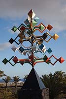 Multi coloured windmill, metal pyramid sculpture by Cesar Manrique on black volcanic rock plinth with sea view and palm trees.
