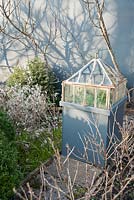 A cast iron Victorian lantern cloche set on a pedestal in a formal kitchen garden surrounded by fruit bushes casting shadows onto the grey wall behind.