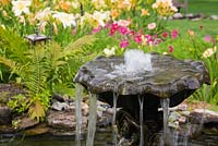 Close-up of cascading water fountain in pond bordered by Pteridophyta - Ferns and colourful palette of Hemerocallis - Daylily and purple Cosmos flowers in backyard Country garden in summer, Jardin des Mesanges garden, Quebec, Canada