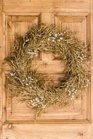 Simple Christmas wreath on front door that blends beautifully with the wood. A wire wreath frame is covered in lengths of golden grass, sprayed gold, and secured in place with florists' wire. It is then punctuated with sprays of quaking grass, sprayed silver.