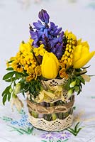 A colourful spring posie of everyday garden flowers - fragrant Mahonia japonica, Clematis cirrhosa var. ballearica, viola, hyacinth, winter jasmine, dwarf golden tulip and heather in a  glass jar decoratd with cotton lace and jute string.