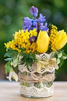 A colourful spring posie of everyday garden flowers - fragrant Mahonia japonica, Clematis cirrhosa var. ballearica, viola, hyacinth, winter jasmine, dwarf golden tulip and heather in a  glass jar decoratd with cotton lace and jute string.