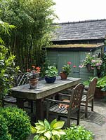 A patio seating area with wooden table and chairs in front of a small shed with green roof.
