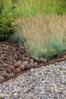 Border containing Festuca glauca 'Blaufuchs' and a mulch of pine cones, beside a path of knapped flint. Garden: The Flintknapper's Garden - A Story of Thetford. Designer: Luke Heydon. Sponsor: Thetford businesses and residents. RHS Hampton Court Flower Show, July 2014