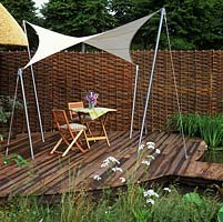 Canvas awning is strung, sail-like, between stainless steel posts to provide shade on a wooden deck. Privacy created from woven willow screens.