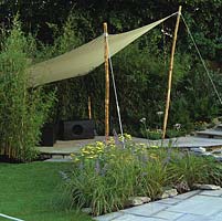 Sail awning on wooden poles by rustling bamboos enclose paved seating area by lily pond, edged in bed planted with achillea, catmint and grasses.