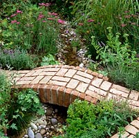 Brick bridge crosses pebble-lined stream edged in achillea, sedum, hardy geranium, alchemilla, lavender, catmint, scabious, mint, sage and parsley.